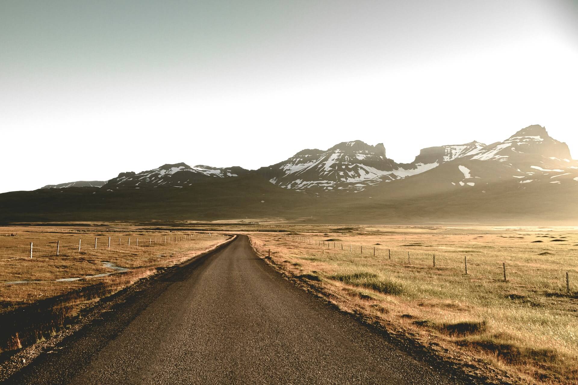 An open road, running past some fields with mountains in the distance